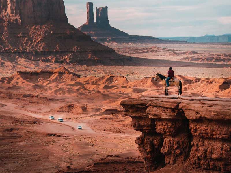 A man riding a horse on a rock ledge, overlooking cars driving through Monument Valley